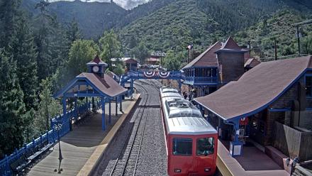 Red cog trains at the Pikes Peak Railway station surrounded by blue trim buildings, tall pines and mountain scenery through changing seasons.