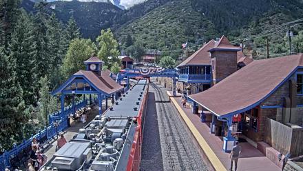 Red cog trains at the Pikes Peak Railway station surrounded by blue trim buildings, tall pines and mountain scenery through changing seasons.