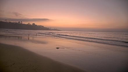 Beach camera over the sand at La Jolla Shores with wide surf and waves, downtown La Jolla visible in the background along the San Diego coastline.