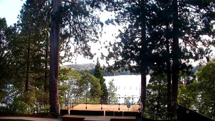 A bright lakeside view from Lake Arrowhead Resort showing green lawn, tall pine trees, and blue lake water beyond.