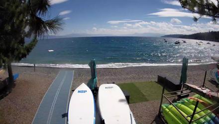 Fixed webcam at Watermans Landing shows paddle boards on a rocky beach, blue Lake Tahoe and Sierra peaks beyond.