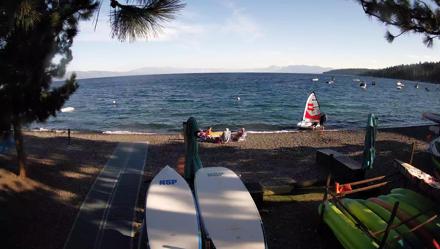 Fixed webcam at Watermans Landing shows paddle boards on a rocky beach, blue Lake Tahoe and Sierra peaks beyond.