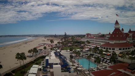 A high resolution view over the Hotel del Coronado resort grounds with pool, beach, and distant coastline under calm skies.
